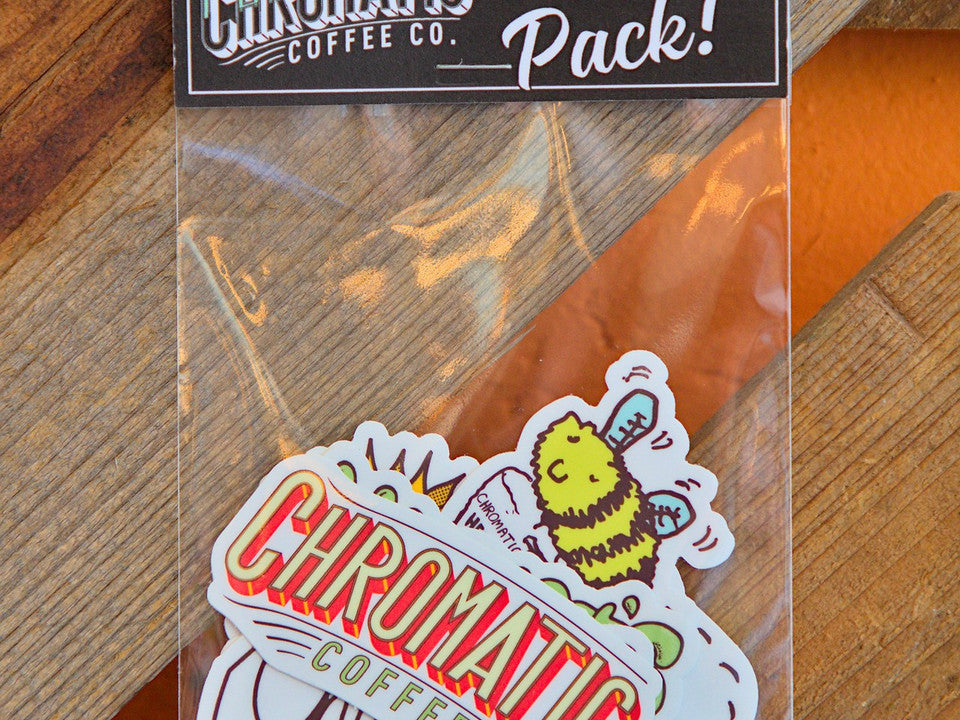 A sticker pack from Chromatic Coffee Co. displayed on a wooden surface, including various stickers with coffee-related illustrations and text.