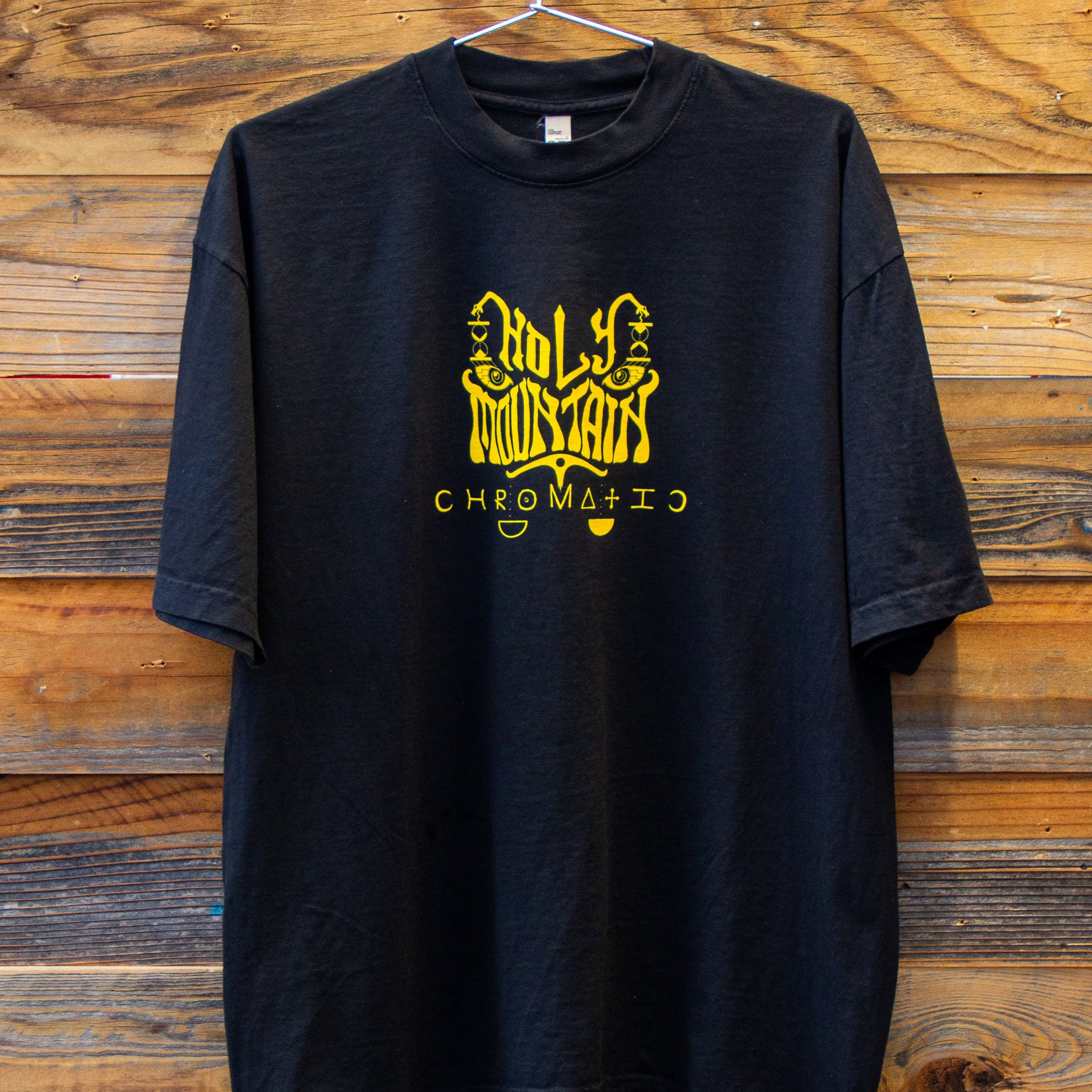 Holy Mountain Shirt Chromatic Coffee Black t shirt with yellow logo on a wooden background