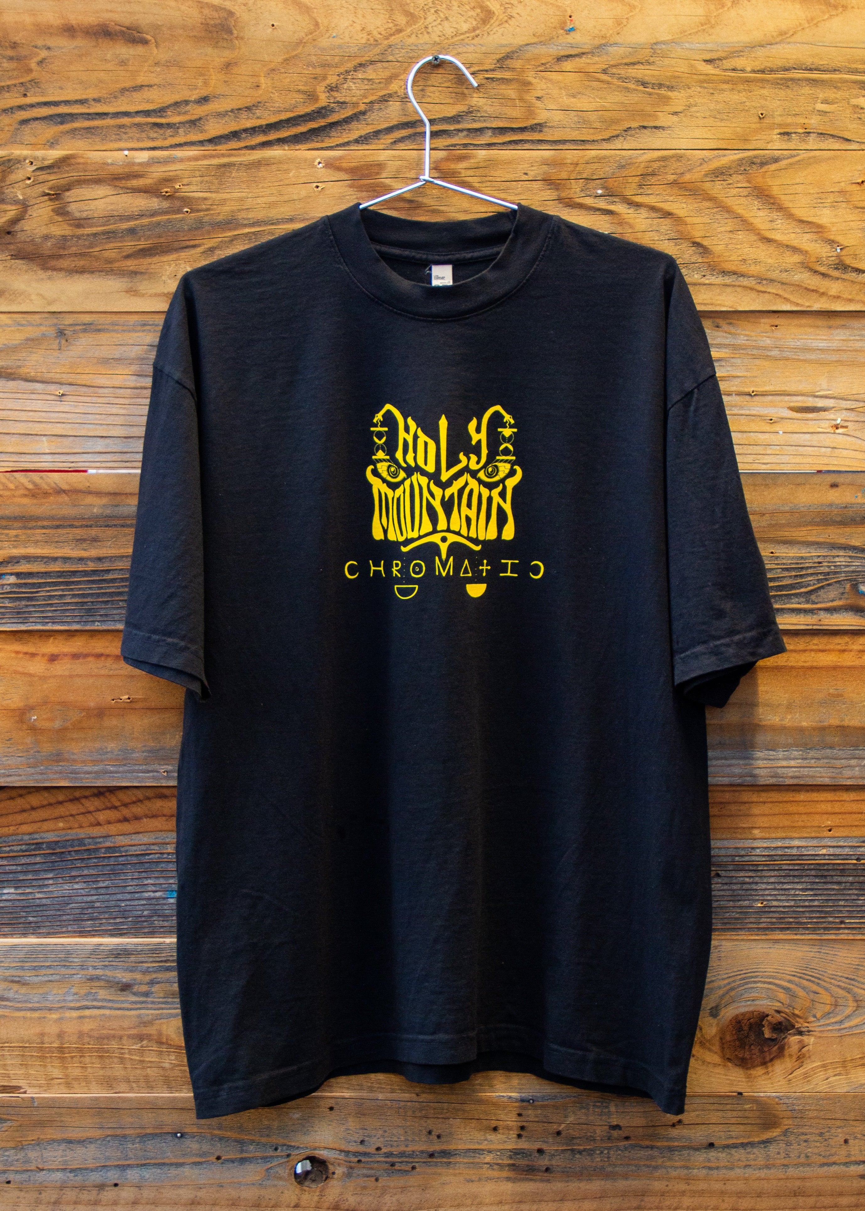 Holy Mountain Shirt Chromatic Coffee Black t shirt with yellow logo on a wooden background