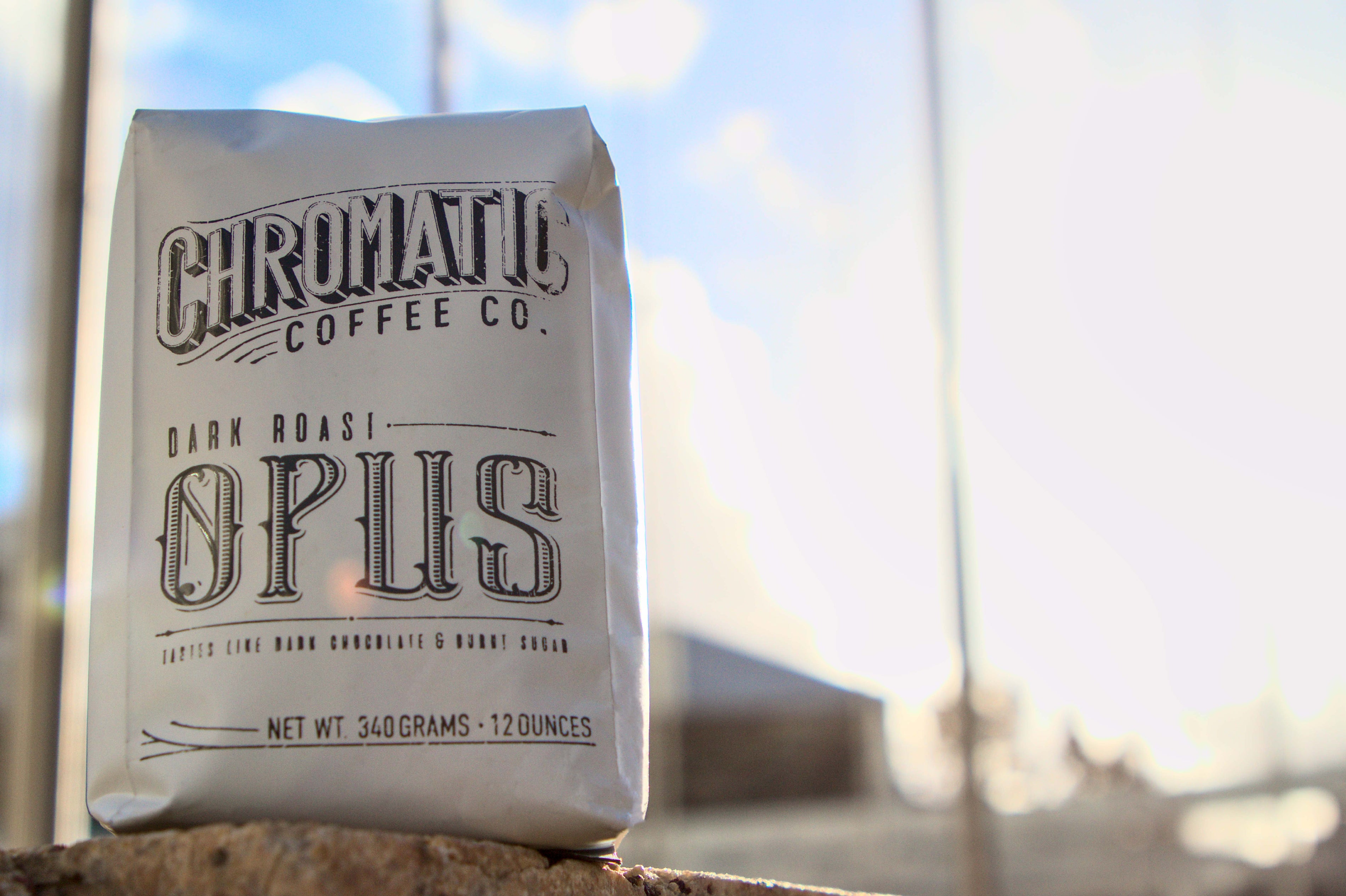 A pack of Opus Dark Roast coffee by Chromatic Coffee Co., standing in front of a window with a cloudy sky in the background.
