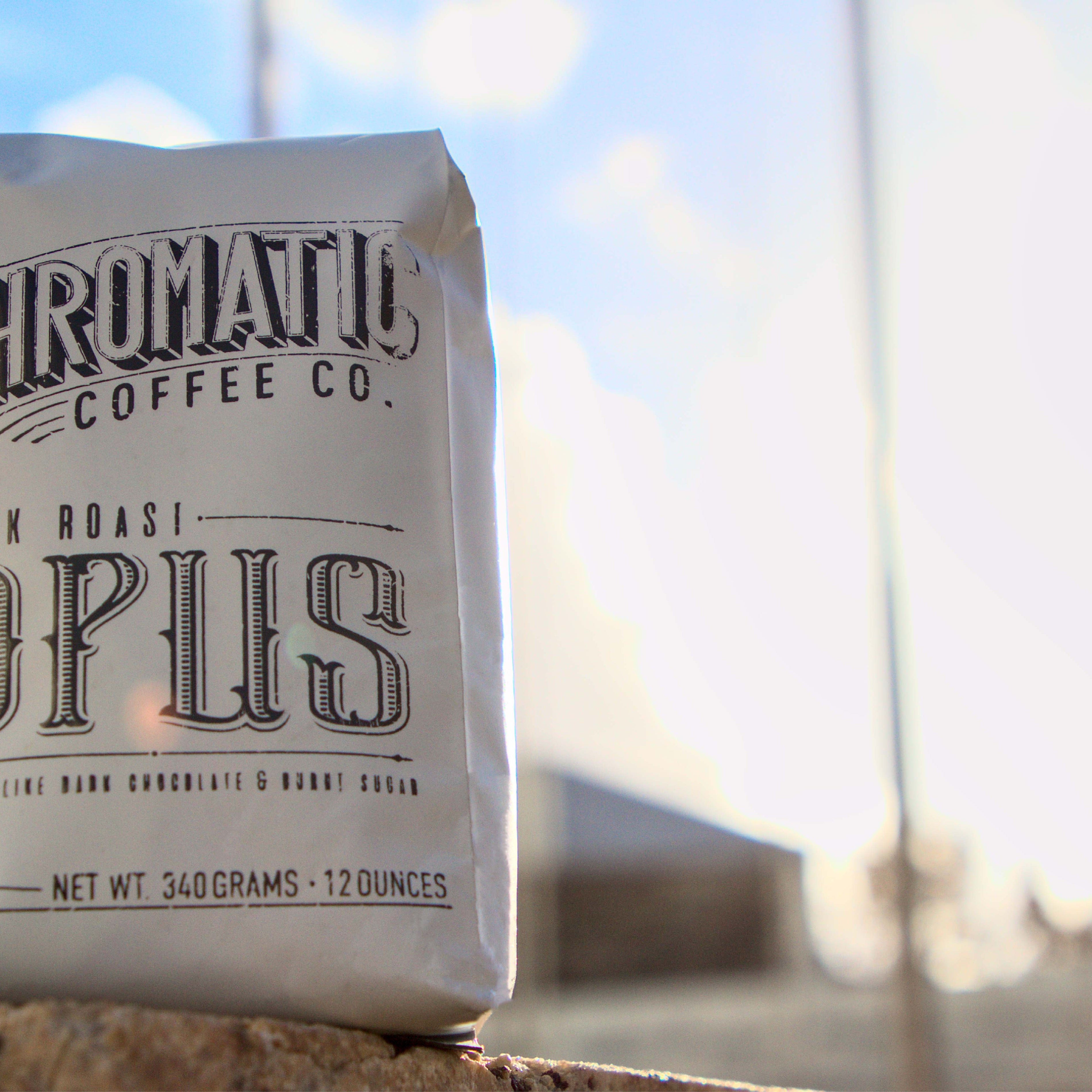 A pack of Opus Dark Roast coffee by Chromatic Coffee Co., standing in front of a window with a cloudy sky in the background.