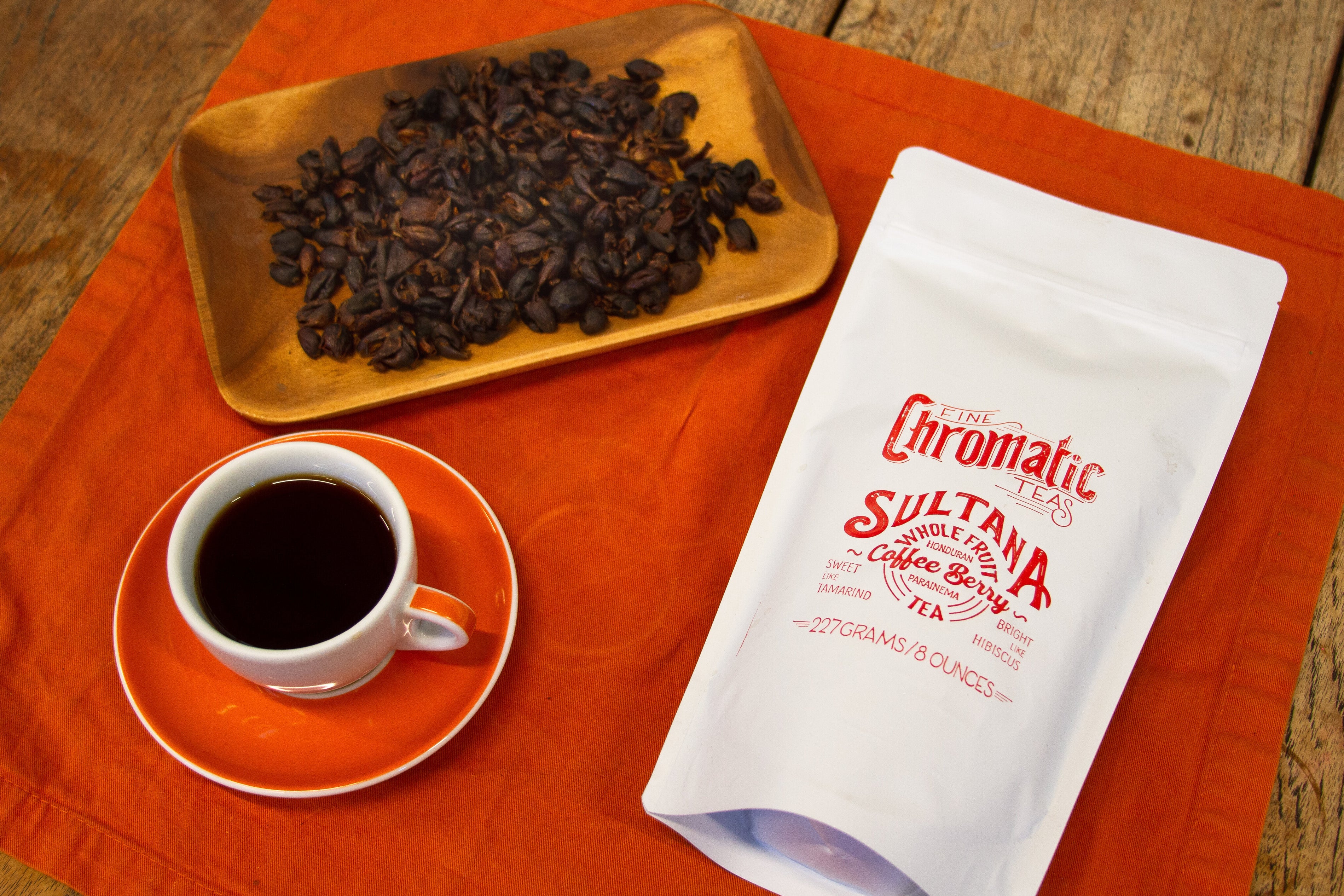 A package of Chromatic Sultana Coffee Berry Tea alongside a cup of tea and a plate of coffee beans on an orange cloth.