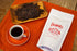 A package of Chromatic Sultana Coffee Berry Tea alongside a cup of tea and a plate of coffee beans on an orange cloth.