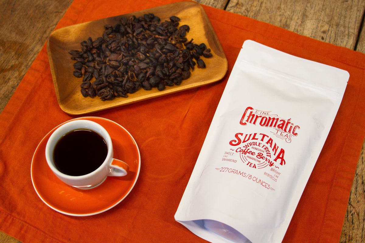 A package of Chromatic Sultana Coffee Berry Tea alongside a cup of tea and a plate of coffee beans on an orange cloth.