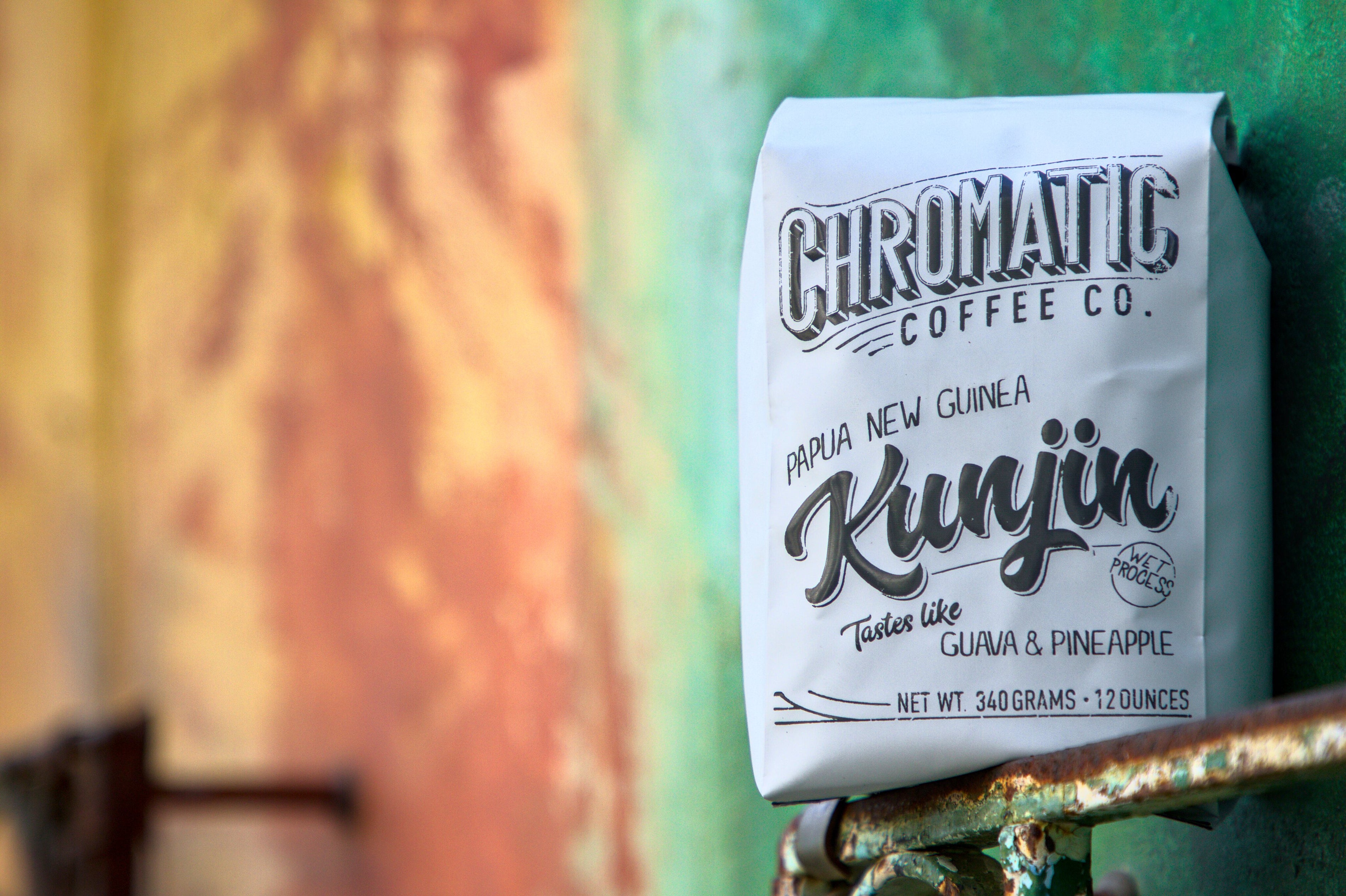 A pack of Chronic Coffee Co. Papua New Guinea Kunjin coffee, with branding and product information, placed on a rusted metal surface against a colorful wall.