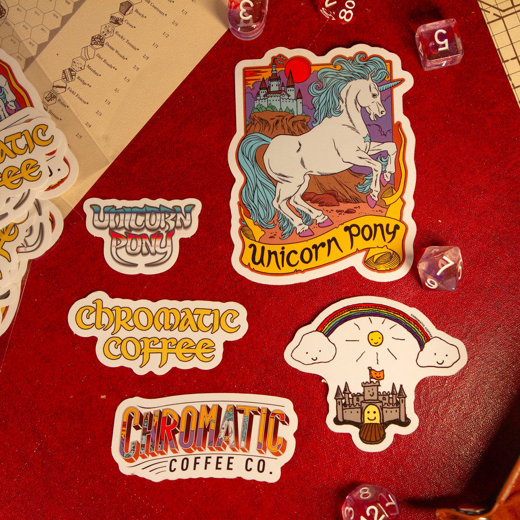 A collection of colorful stickers with unicorn and castle illustrations, branding for Chromatic Coffee, and other related designs laid out on a red surface.